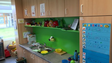 Einzeilige Küche in einem Kindergartencontainer mit hellbraunen Fronten, grauer Arbeitsplatte und grünem Wandspiegel