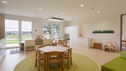 Gruppenraum in einem Kindergarten-Modulbau mit kindgerechter Möblierung, Küchenzeile, Doppelflügel-Terrassentüren, Akustikdecke und grünen Dekoelementen