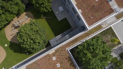 Drohnenaufnahme eines Teilausschnitts eines dreigeschossigen Schulcontainer-Modulgebäudes mit Holzfassade, Gründach, Spiel- und Grünflächen, genutzt als Kinderhort