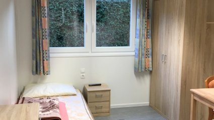 Einzelzimmer in einem Wohncontainer, ausgestattet mit einem Bett, Kasten, Kommode, Tisch, einem Doppelfenster und Vorhängen
