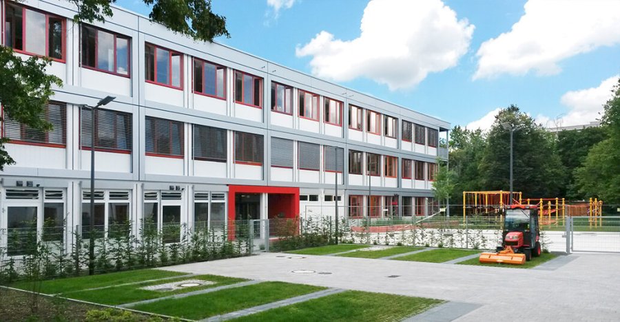 Dreistöckiges Schulcontainer-Gebäude mit rotem Eingangsbereich, grauer Alucobond-Geschoßverblendung, Fenstern mit roten Rahmen, vorgelagertem Parkplatz und Schulhof