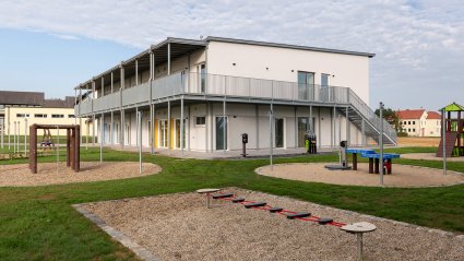 Seitliche Außenansicht eines zweistöckigen Kindergarten-Modulbaus mit überdachter Balkonanlage, Fluchttreppe und Spielplatzanlage