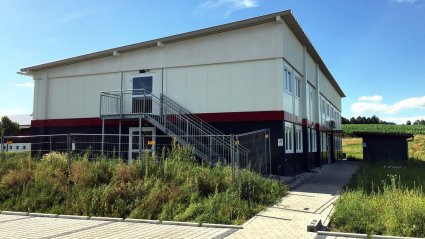 Seitenansicht eines Kindergarten-Modulbaus mit dunkelgrauen Modulen im Erdgeschoß, weißen Modulen im Obergeschoß, roter Geschoßverblendung, Fluchttreppe und Pultdach
