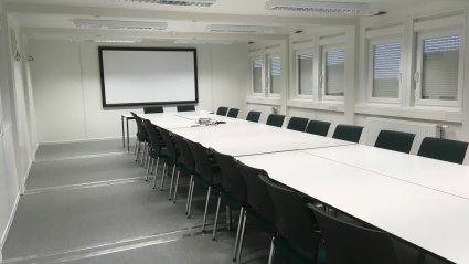 Gro&szlig;er Besprechungsraum in einem Containergeb&auml;ude mit Besprechungstischen, schwarzen St&uuml;hlen, Projektionsfl&auml;che und Fenstern mit Rolll&auml;den
