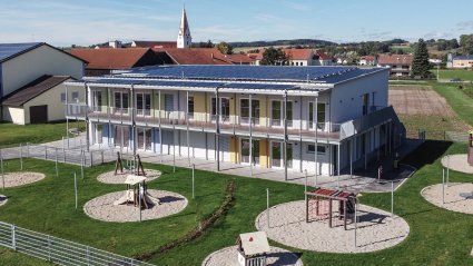 Drohnenaufnahme eines zweistöckigen Kindergarten-Modulbaus mit überdachter Balkonanlage, Fluchttreppe, Flachdach, PV-Analage und Spielplatz