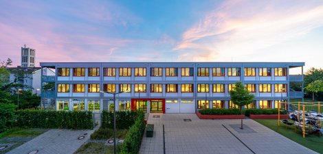 Frontalansicht eines dreigeschoßigen Schulcontainer-Modulbaus mit roten Fenstern, grauen Containern und grauer Alucobond-Geschoßverkleidung