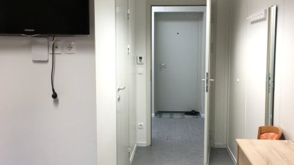 Zimmerflur in einem Wohncontainer-Modulbau mit offener Tür zum Gang, Garderobenbereich, Kommode, Sessel und Fernseher