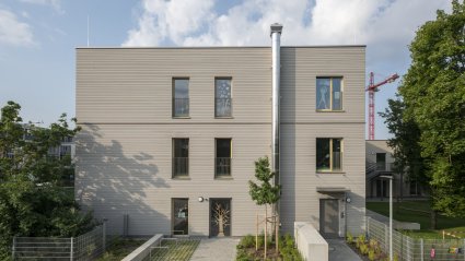 Seitliche Außenaufnahme eines dreigeschoßigen Schulcontainer-Modulbaus mit Holzfassade, außenliegendem Edelstahl-Kaminrohr, gepflastertem Gehweg und Fahrradabstellplätzen