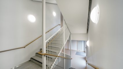 Innenaufnahme des Treppenhauses eines Schulcontainer-Modulbaus mit Betonstufen, schwarzem Kantenschutz, weißem Stahlgeländer mit Holzhandlauf, und runden Wandleuchten