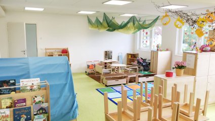 Gruppenraum in einem Kindergarten-Modulbau mit kindgerechtem Mobiliar, grünem Vinylboden, weißen Wänden und Doppelfenstern