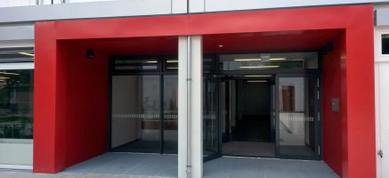 Mehrstöckiger Modulbau mit großzügigen Eingangsbereich, verkleidet mit rotem Alucobond, zwei Doppelflügel-Eingangstüren und Abstreif-Gitterrost