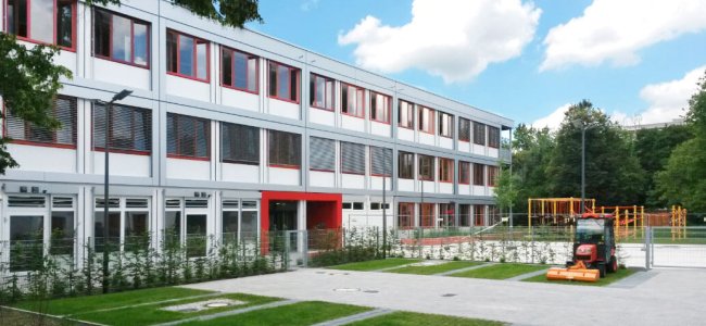 Modulbau - Schulcontainer Dreigeschossiger Schulcontainer-Modulbau mit grauer Alucobond Etagen- und Geschoß-Verkleidung, rote Fensterrahmen, roter Eingangsbereich, Parkplatz und Spielplatz