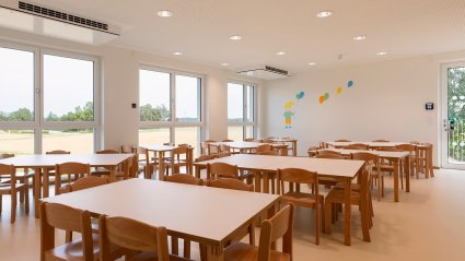 Speisesaal in einem Kindergarten-Modulbau mit kindgerechten Tischen und Stühlen, Doppelfenstern mit bodenebener Fixverglasung und Notausgangstür