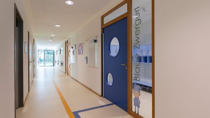 Gang in einem Kindergarten-Modulbau mit hellem Boden und weißen Wänden, buntem Bodenleitsystem zur gelben und blauen Gruppentür