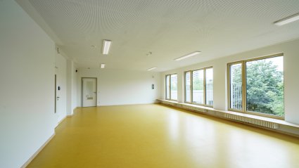 Innenaufnahme eines großen Gruppenraums in einem Schulcontainer-Modulgebäude mit gelbem Vinylboden, Holz-Sockelleisten, Fensterfront mit vorgebauter Sitzgelegenheit, weißen Wänden und Akustikdecke