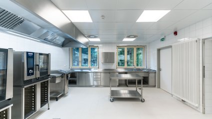 Innenaufnahme einer Küche in einem Schulcontainer-Modulbau mit Edelstahl-Küchengeräten, Edelstahl-Dunstabzug, weißen Wandfliesen, Bodenabfluss, zwei Dreiflügel-Fenster