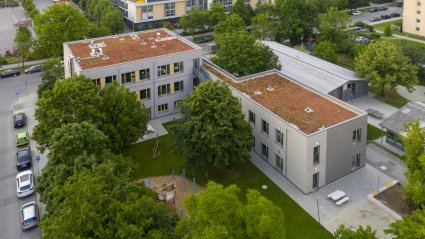 Drohneaufnahme eines dreistöckigen Schulcontainer-Modulgebäudes in L-Form mit Holz-Fassade, Gründach, Spielplatz und Grünflächen