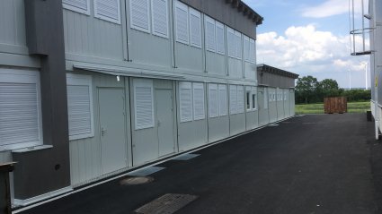 Containergeb&auml;ude aus wei&szlig;en Containern mit Glas&uuml;berdachungen &uuml;ber den Eingangst&uuml;ren, Pultdach und Attika mit Holzverkleidung