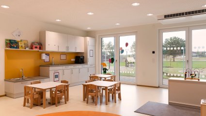 Gruppenraum in einem Kindergarten-Modulbau mit kindgerechter Möblierung, Küchenzeile, Doppelflügel-Terrassentüren, Akustikdecke und orangen Dekoelementen