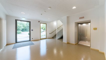 Innenaufnahme des Nebeneingangsbereichs eines Schulcontainer-Modulbaus mit großzügiger Eingangshalle, Stiegenhaus und Aufzug mit geöffneter Lifttür