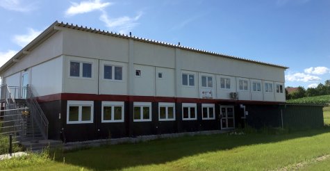 Außenanaufnahme eines Kindergarten-Modulbaus als Kindertagesstätte mit dunkelgrauen Modulen im Erdgeschoß, weißen Modulen im Obergeschoß, roter Geschoßverblendung, Fluchttreppe und Pultdach