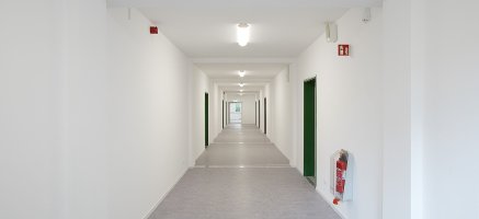 Heller Flur in modularem Gebäude mit grauen Vinylboden, grünen Türen, Feuerlöscher und Beleuchtung