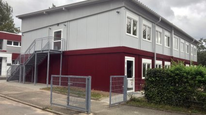 Zweistöckige Schulcontaineranlage mit rot lackierten Containern im Erdgeschoss, grau lackierten Containern im Obergeschoss, Stahl-Außentreppe, Pultdach und Wellblechdach