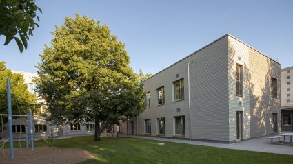 Außenaufnahme einer zweigeschoßigen Schulcontaineranlage für einen Kinderhort mit Holzverkleidung und vorgelagertem Spielplatz mit Grünfläche