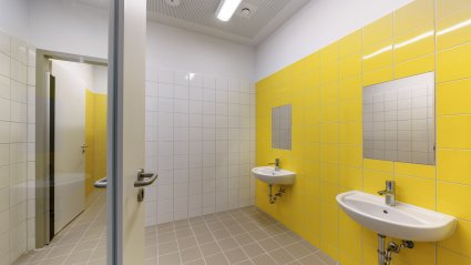 Innenansicht des Sanitärbereichs eines Schulcontainer-Modulbaus mit hellbraunen Bodenfliesen, weißen und gelben Wandfliesen, zwei Waschbecken mit Spiegel und separatem Zugang zum WC-Bereich