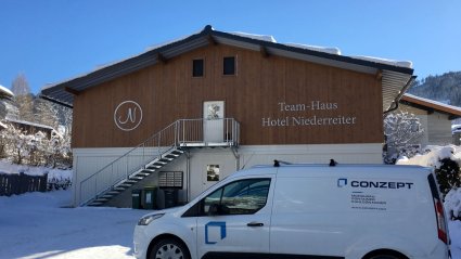 Vorderansicht Eingangsbereich einer Wohncontainer-Anlage mit weißen Modulen im Erdgeschoß, Holzfassade im Obergeschoß, Außentreppe und Satteldach