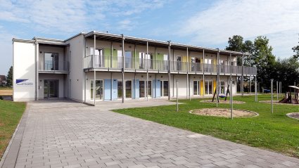 Außenansicht eines zweistöckigen Kindergarten-Modulbaus mit überdachter Balkonanlage, Spielplatz und gepflastertem Gehweg