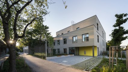 Außenaufnahme des Eingangsbereichs mit gelben Wandelementen und großem Vorplatz eines dreigeschoßigen Schulcontainer-Modulgebäudes für einen Kinderhort mit Holzfassade