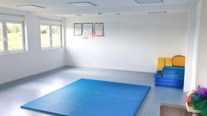 Bewegungsraum in einem Kindergarten-Modulbau mit hellblauem Boden, weißen Wänden, Doppelfenstern und blauen Turnmatten