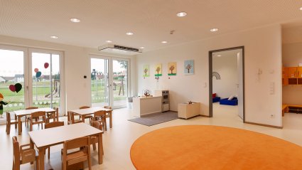 Gruppenraum in einem Kindergarten-Modulbau mit kindgerechter Möblierung, Doppelflügel-Terrassentüren, Verbindungstür zum Schlafraum und orangen Dekoelementen