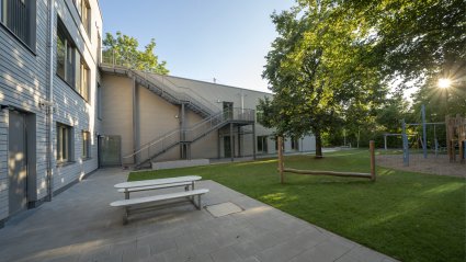 Schulcontainer-Modulgebäude mit Holzfassade und Nottreppe, vorgelagerte Terrasse mit Sitzgelegenheit und Tisch, Spielplatz und Grünfläche