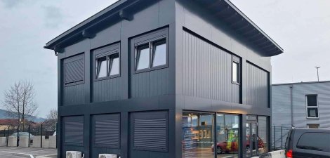 Seitliche Vorderansicht eines modernen zweigeschossigen Containerb&uuml;ros mit integriertem Wohncontainer in Anthrazit, Pultdach und vollfl&auml;chiger Verglasung &uuml;ber die gesamte L&auml;nge des Erdgeschosses